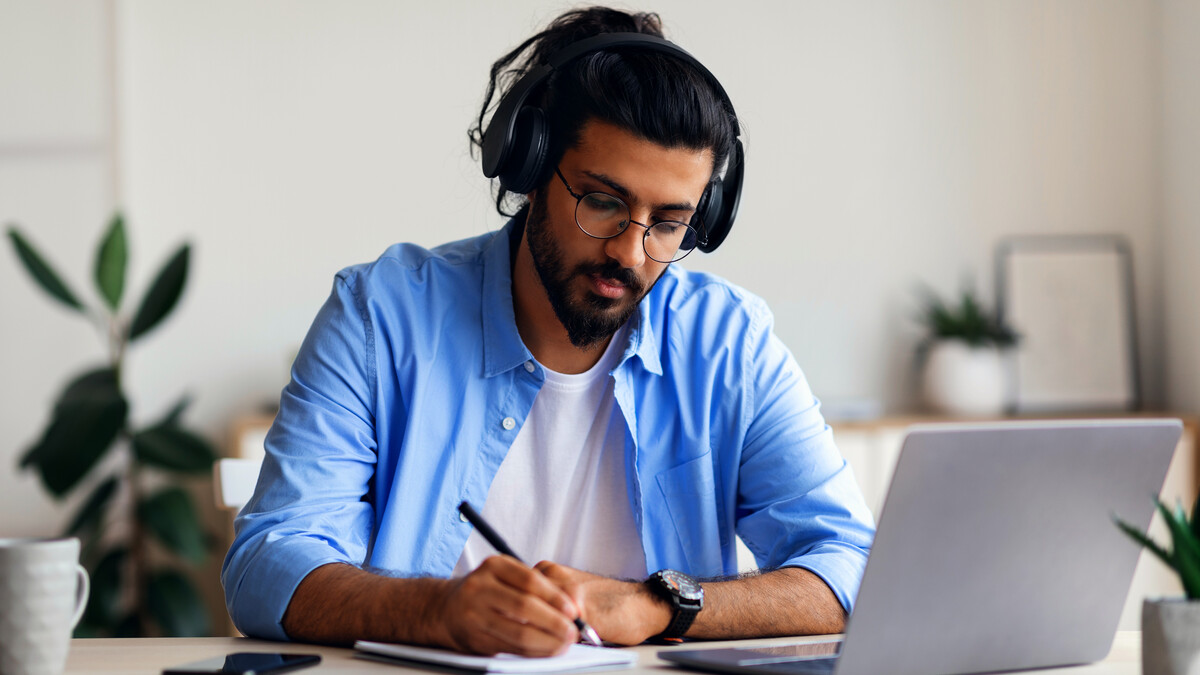 Professional wearing headphones and taking notes beside a laptop.