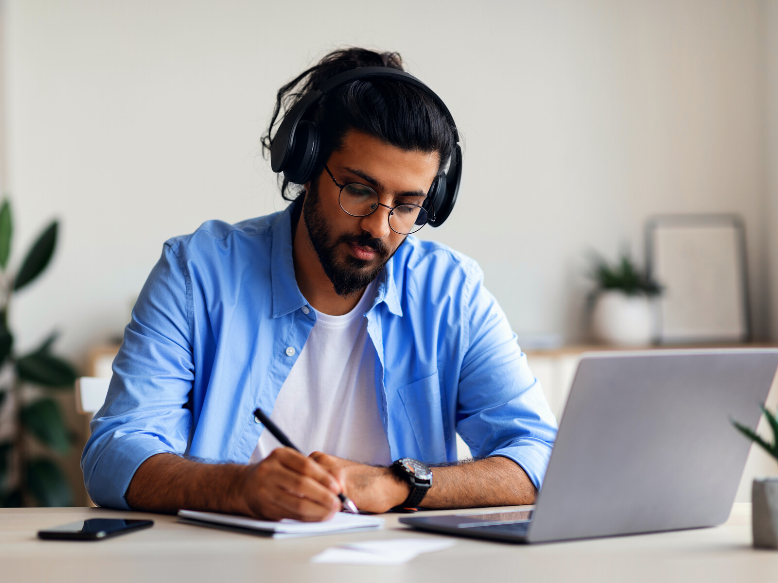 Professional wearing headphones and taking notes beside a laptop.